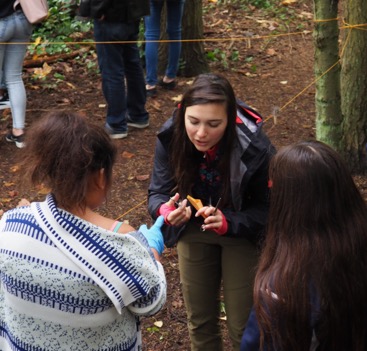 Lauren Henson, PhD student in the Raincoast Applied Conservation Science lab standing in the forest with students near the hair snag. She is holding forceps and an envelope to demonstrate hair collection methods to the students surrounding her.