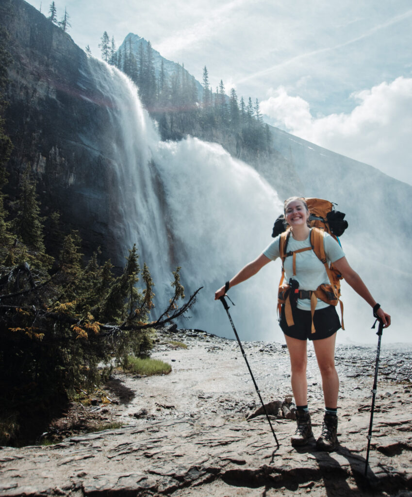 Emma Bialobzyski smiles brightly, carrying a large backpack and hiking poles. In the background, a roaring waterfall cascades down the side of a mountain, the sun is shining.