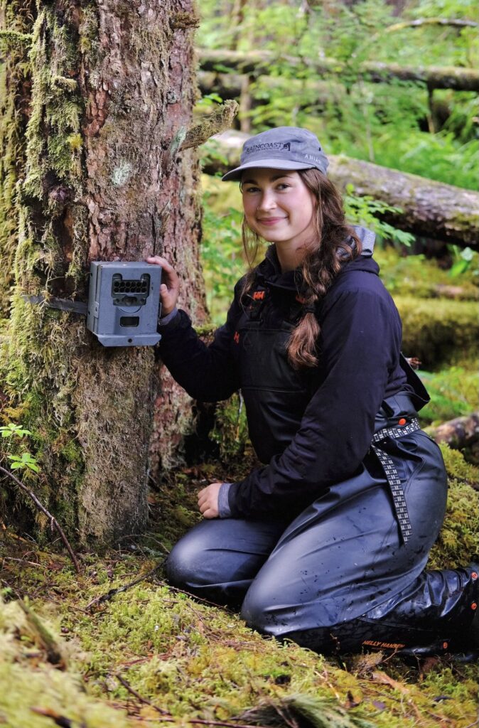 Abby Tinsley kneels beside a tree, smiling, setting up a remote wildlife camera on the Central Coast of British Columbia, surrounded by moss and trees.